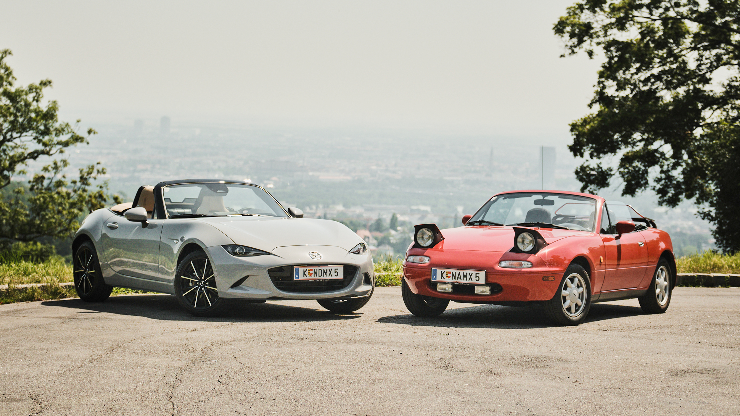 Ein altes, rotes Cabrio und ein modernes, graues Cabrio stehen auf einem Parkplatz, im Hintergrund ist eine Stadt zu sehen.