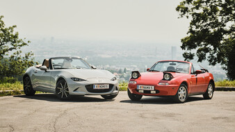 Ein altes, rotes Cabrio und ein modernes, graues Cabrio stehen auf einem Parkplatz, im Hintergrund ist eine Stadt zu sehen.