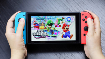 Nintendo Switch Konsole mit blauem und roten Controller im Handheldmodus. Person hält die Konsole und spielt das Videospiel Mario Bros. Wonder.