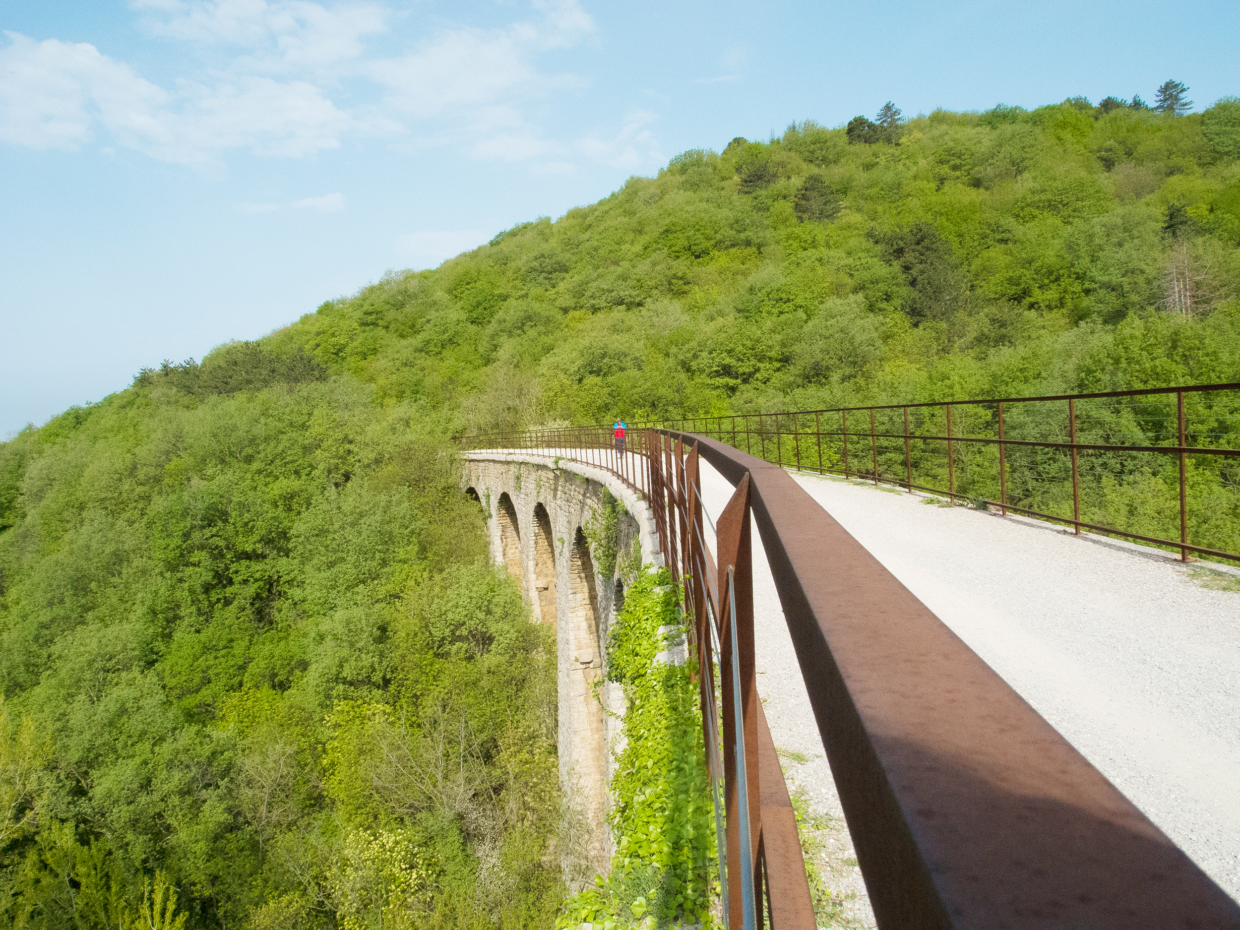 Ein Radfahrer überquert ein großes Viadukt in einer mit Bäumen bedeckten hügeligen Landschaft. Der Radweg ist Teil einer stillgelegten Bahnstrecke von Triest hinauf in den Karst. 