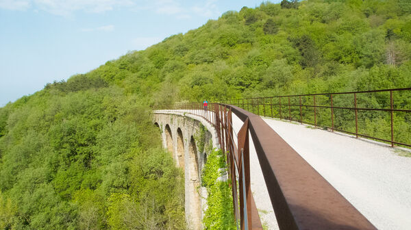 Ein Radfahrer überquert ein großes Viadukt in einer mit Bäumen bedeckten hügeligen Landschaft. Der Radweg ist Teil einer stillgelegten Bahnstrecke von Triest hinauf in den Karst.