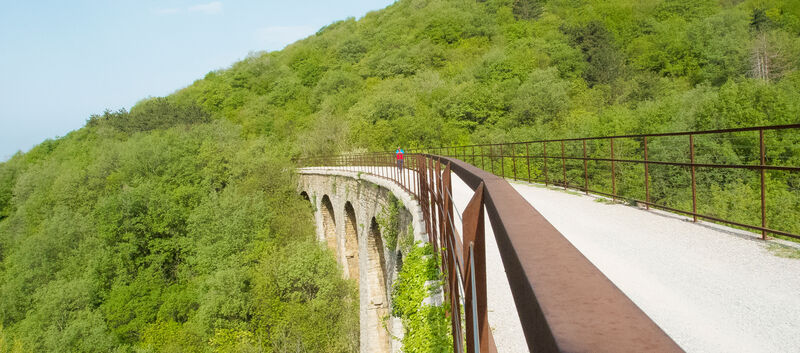 Ein Radfahrer überquert ein großes Viadukt in einer mit Bäumen bedeckten hügeligen Landschaft. Der Radweg ist Teil einer stillgelegten Bahnstrecke von Triest hinauf in den Karst.