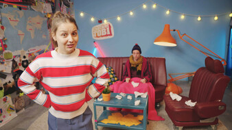 Checkerin Marina im Vordergrund im Checker-Studio. Im Hintergrund sitzt Checker Tobi mit einer Decke am Sofa.
