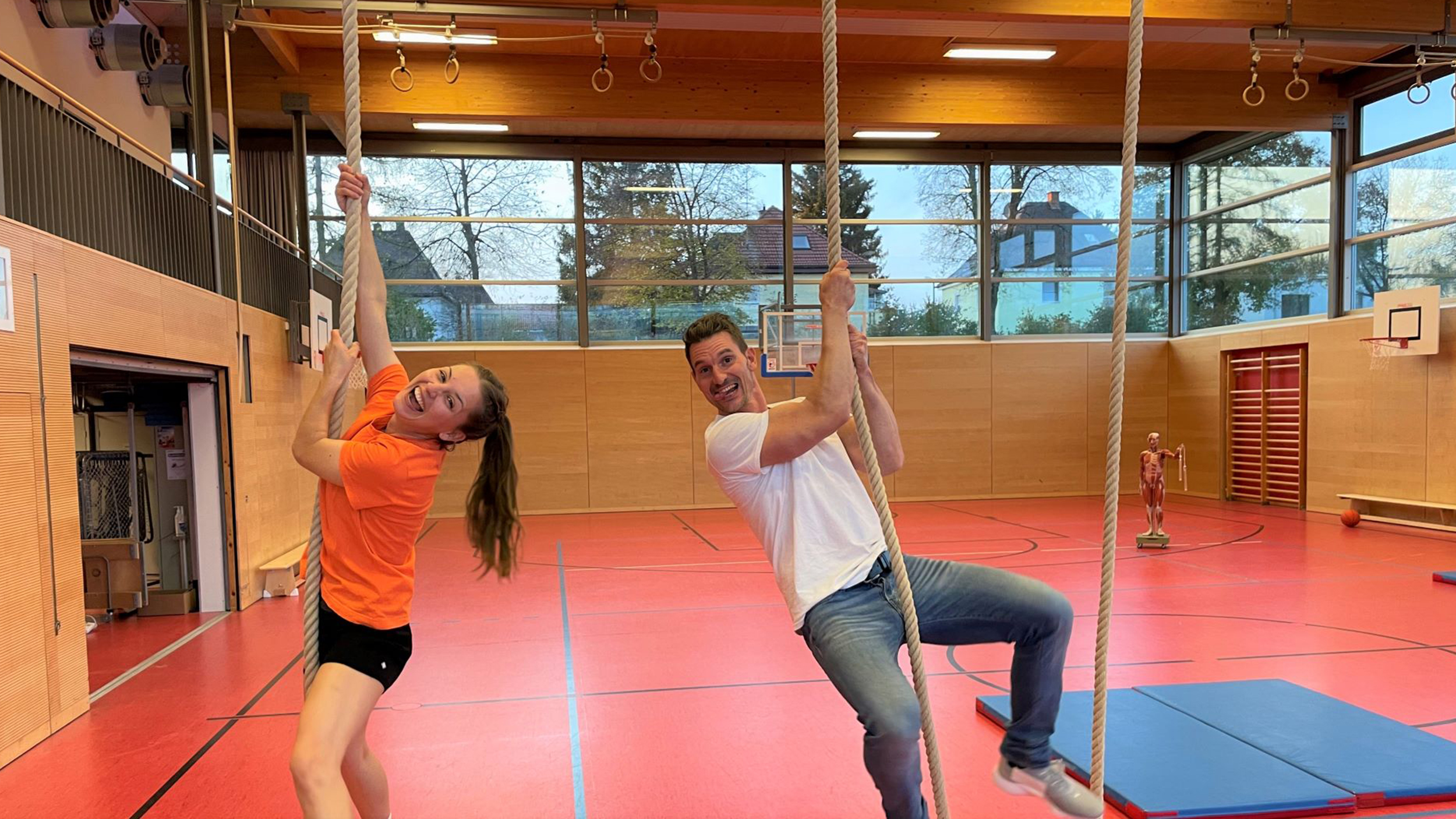 Checkerin Marina und Trainer klammern sich an Seilen fest in einem großen Turnsaal.