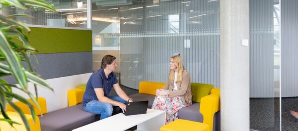 Im Bürobereich. Im Vordergrund links eine Pflanze. in der Mitte eine gelb-graue Couch auf der zwei Personen sitzen. Ein Mann mit dunklem T-Shirt und Jeans und eine Frau in einem gemusterten Kleid. Sie reden miteinander. Vor ihnen, auf einem weißen Couchtisch, steht ein Laptop. Im Hintergrund geht eine Frau mit rosa Hose und weißem Top an ihnen vorbei.