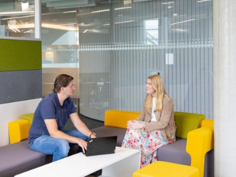 6.jpg Im Bürobereich. Im Vordergrund links eine Pflanze. in der Mitte eine gelb-graue Couch auf der zwei Personen sitzen. Ein Mann mit dunklem T-Shirt und Jeans und eine Frau in einem gemusterten Kleid. Sie reden miteinander. Vor ihnen, auf einem weißen Couchtisch, steht ein Laptop. Im Hintergrund geht eine Frau mit rosa Hose und weißem Top an ihnen vorbei.