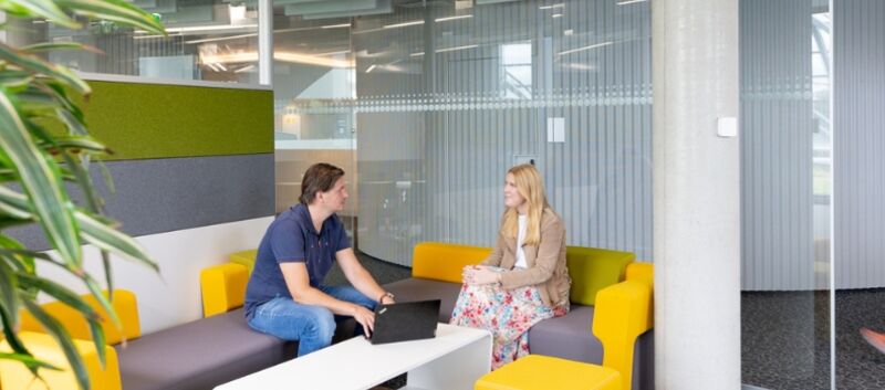 Im Bürobereich. Im Vordergrund links eine Pflanze. in der Mitte eine gelb-graue Couch auf der zwei Personen sitzen. Ein Mann mit dunklem T-Shirt und Jeans und eine Frau in einem gemusterten Kleid. Sie reden miteinander. Vor ihnen, auf einem weißen Couchtisch, steht ein Laptop. Im Hintergrund geht eine Frau mit rosa Hose und weißem Top an ihnen vorbei.