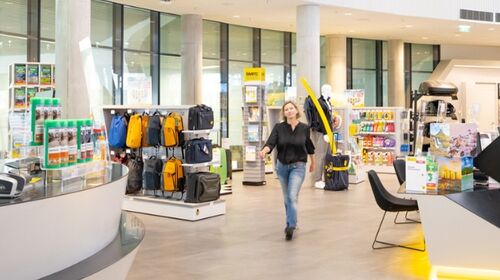 3.jpg Im Schalterbereich eines ÖAMTC Stützpunktes. Eine Person schlendert auf die Kamera zu. Die Frau trägt ein schwarzes Top und eine Jeans und hat schulterlanges, blondes Haar. Rechts von ihr befinden sich Produkt-Regale, links von ihr der Wartebereich für Kundinnen.