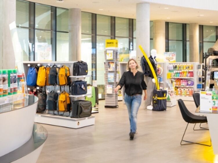 Im Schalterbereich eines ÖAMTC Stützpunktes. Eine Person schlendert auf die Kamera zu. Die Frau trägt ein schwarzes Top und eine Jeans und hat schulterlanges, blondes Haar. Rechts von ihr befinden sich Produkt-Regale, links von ihr der Wartebereich für Kundinnen.