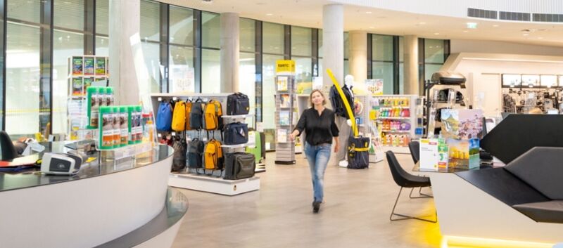 Im Schalterbereich eines ÖAMTC Stützpunktes. Eine Person schlendert auf die Kamera zu. Die Frau trägt ein schwarzes Top und eine Jeans und hat schulterlanges, blondes Haar. Rechts von ihr befinden sich Produkt-Regale, links von ihr der Wartebereich für Kundinnen.