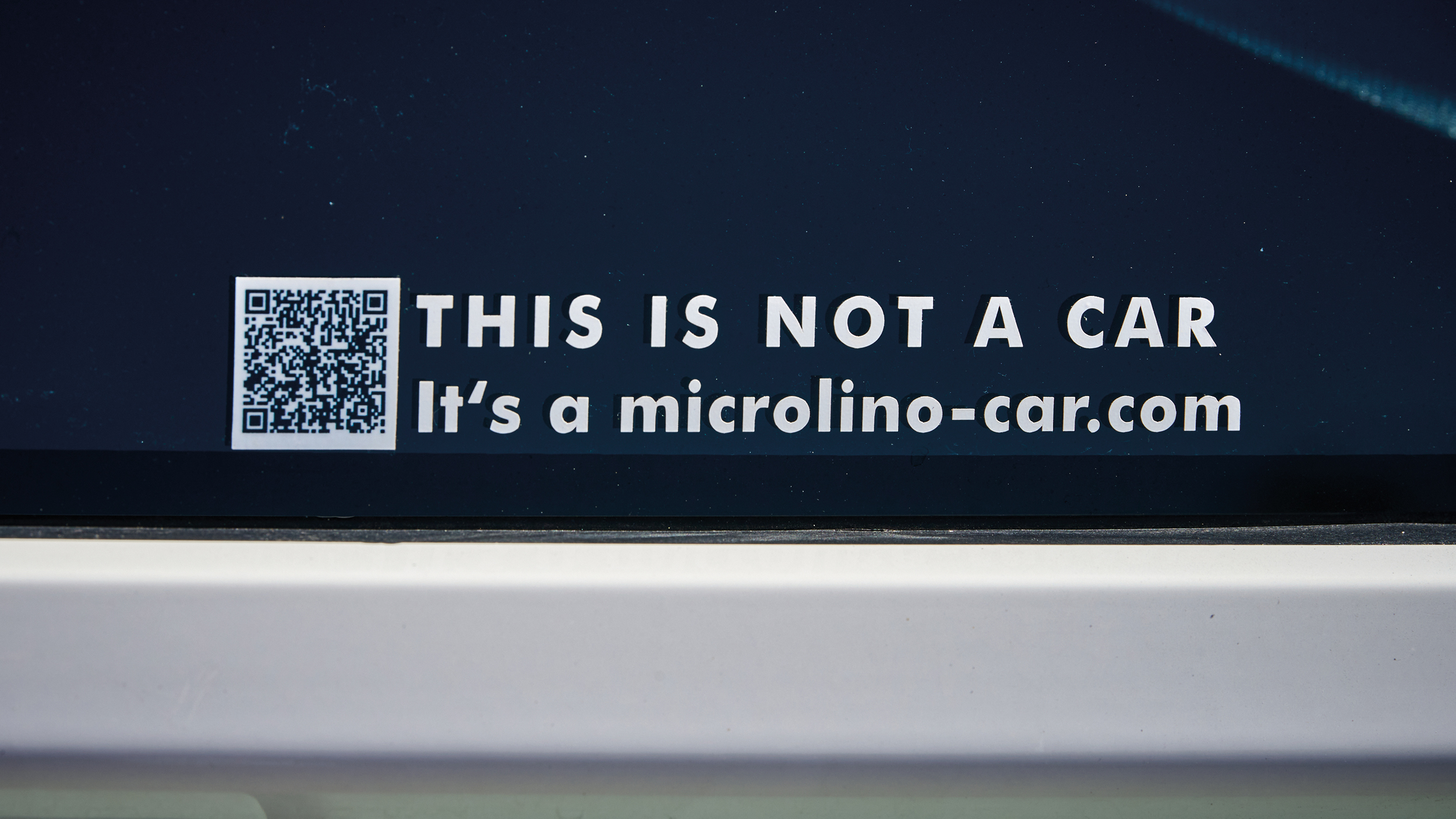 Ein Aufkleber mit QR-Code, der "This is not a car. It's a microlino-car.com"  besagt, auf dem Seitenfenster des Kleinfahrzeuges Microlino, 