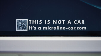 Ein Aufkleber mit QR-Code, der "This is not a car. It's a microlino-car.com" besagt, auf dem Seitenfenster des Kleinfahrzeuges Microlino,