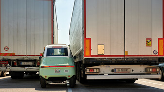 Das hellgrüne Kleinfahrzeug Microlino steht zwischen zwei Lkw.