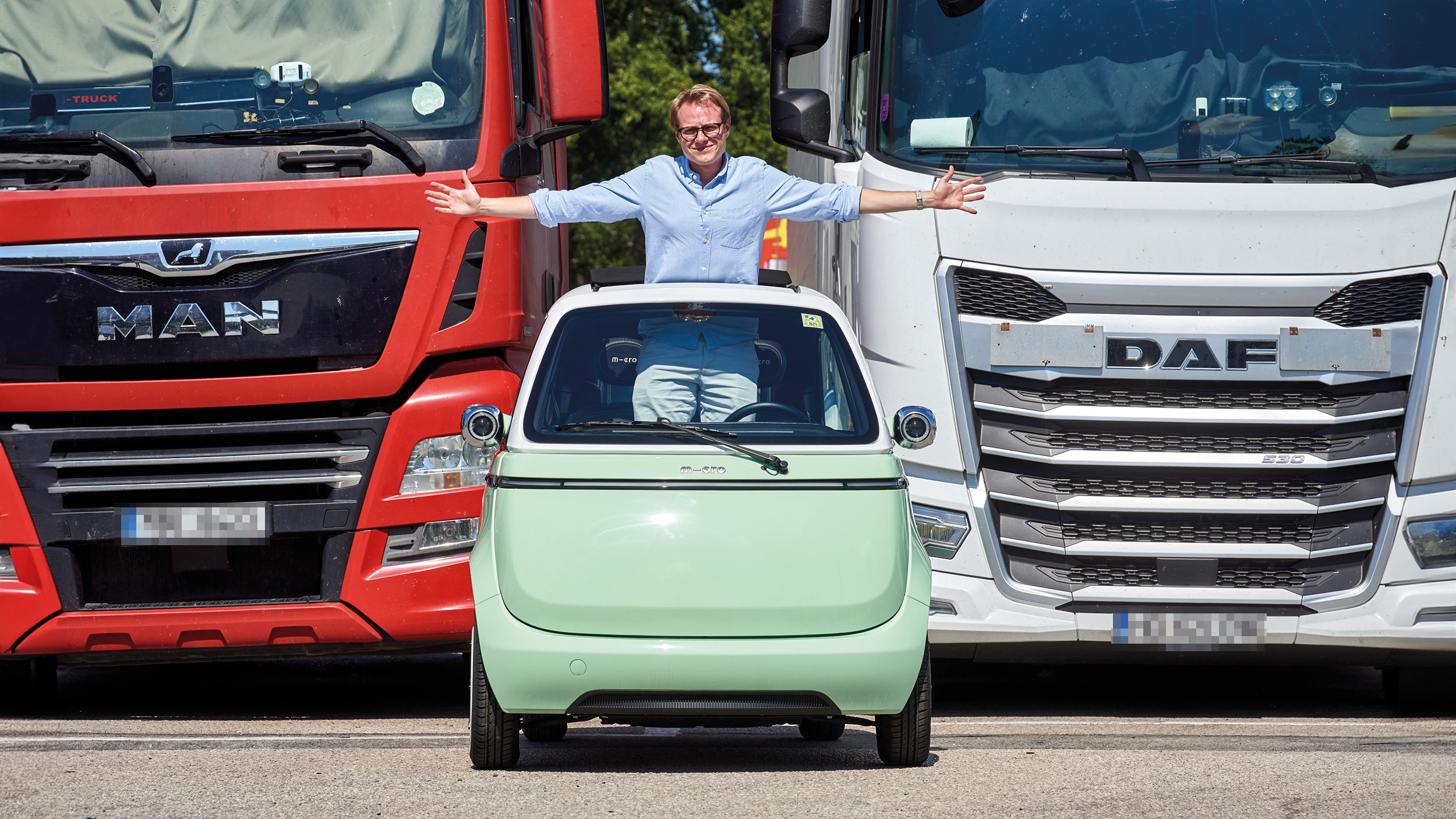 Eine Person steht aufrecht und mit ausgestreckten Armen im hellgrünen Kleinfahrzeug Microlino und blickt durch das Schiebedach. Dahinter stehen ein roter und weißer Lkw.