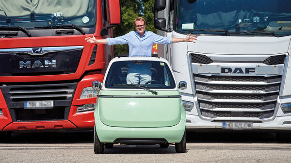 Eine Person steht aufrecht und mit ausgestreckten Armen im hellgrünen Kleinfahrzeug Microlino und blickt durch das Schiebedach. Dahinter stehen ein roter und weißer Lkw.