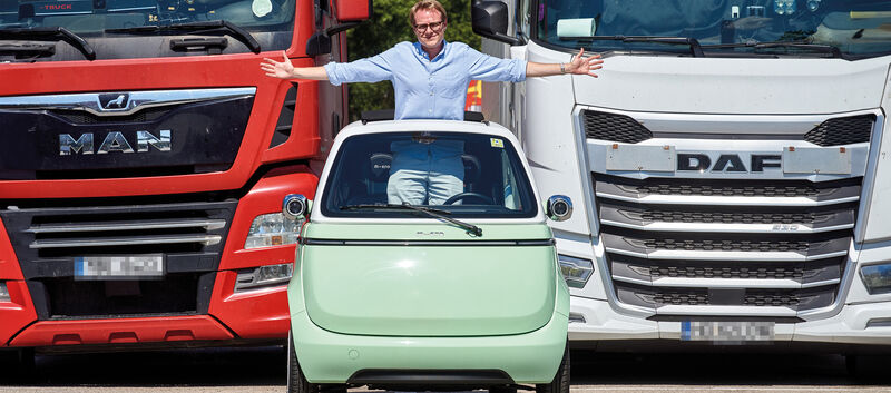 Eine Person steht aufrecht und mit ausgestreckten Armen im hellgrünen Kleinfahrzeug Microlino und blickt durch das Schiebedach. Dahinter stehen ein roter und weißer Lkw.