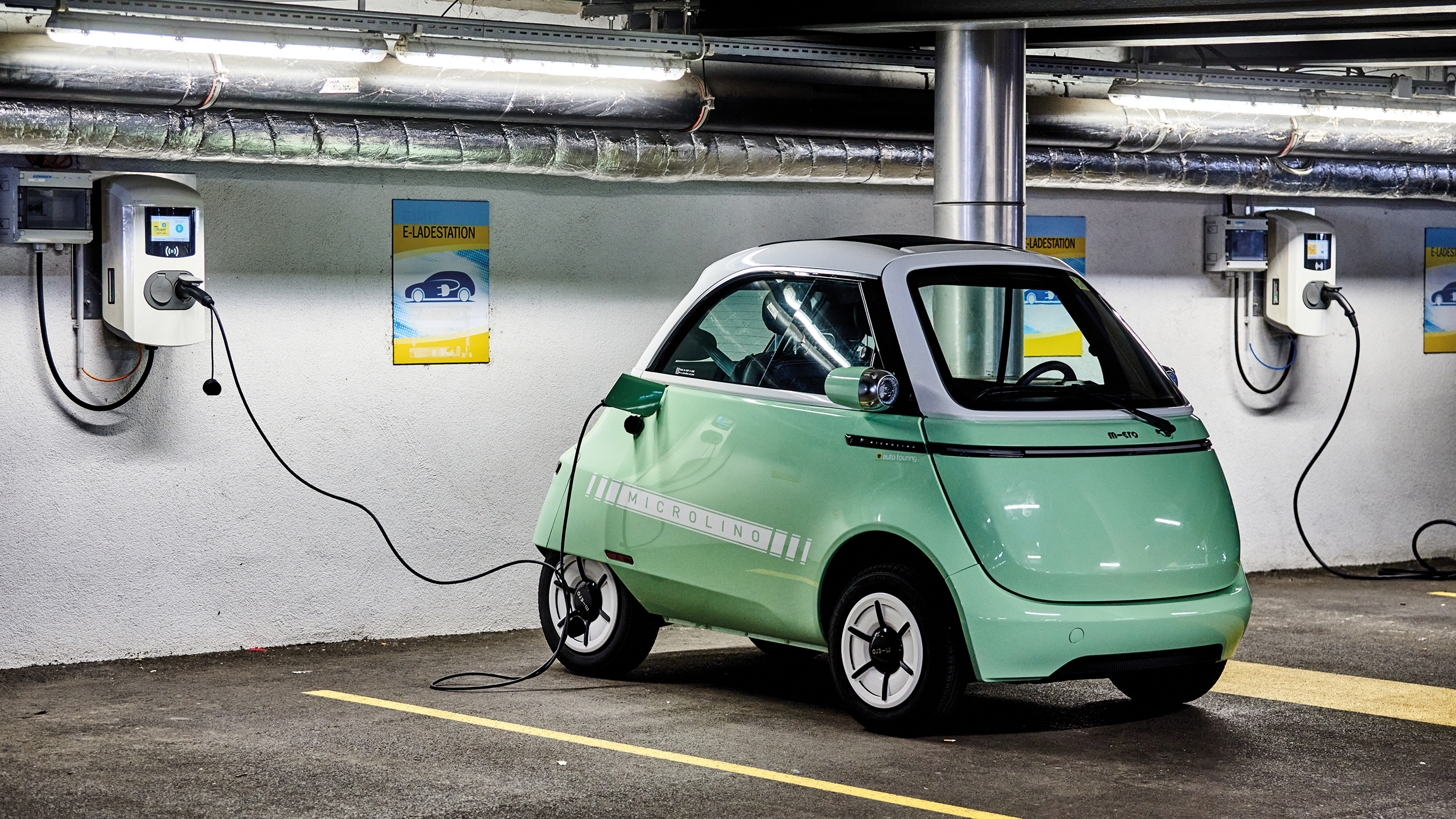 Das Kleinfahrzeug Microlino in hellgrün steht an einer Ladestation angesteckt in einer Garage.