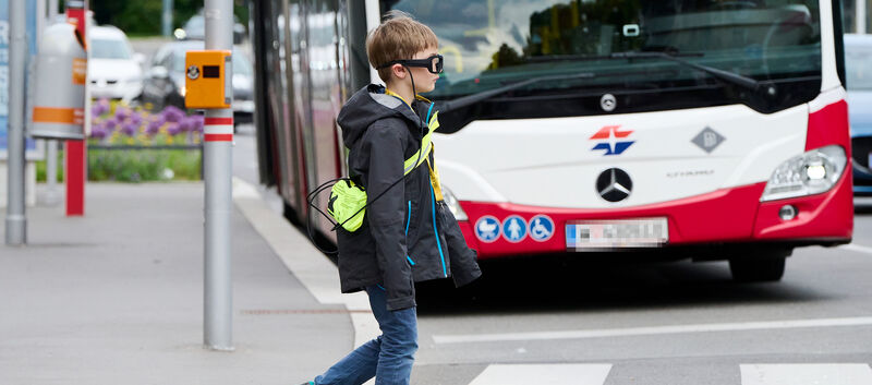 Ein Kind mit einer Blickbewegungsbrille überquert die Straße. Im Hintergrund sieht man einen Autobus.