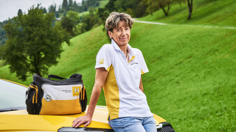 Hauskrankenpflegerin Heidi lehnt auf der Motorhaube ihres Autos. Ihre Tasche steht darauf.