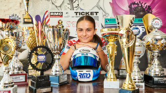 Eine junge Rennfahrerin sitzt auf der Couch und lehnt sich mit ihrem Kinn an einen blauen Rennhelm an, der auf einem Couchtisch voller Pokale steht.