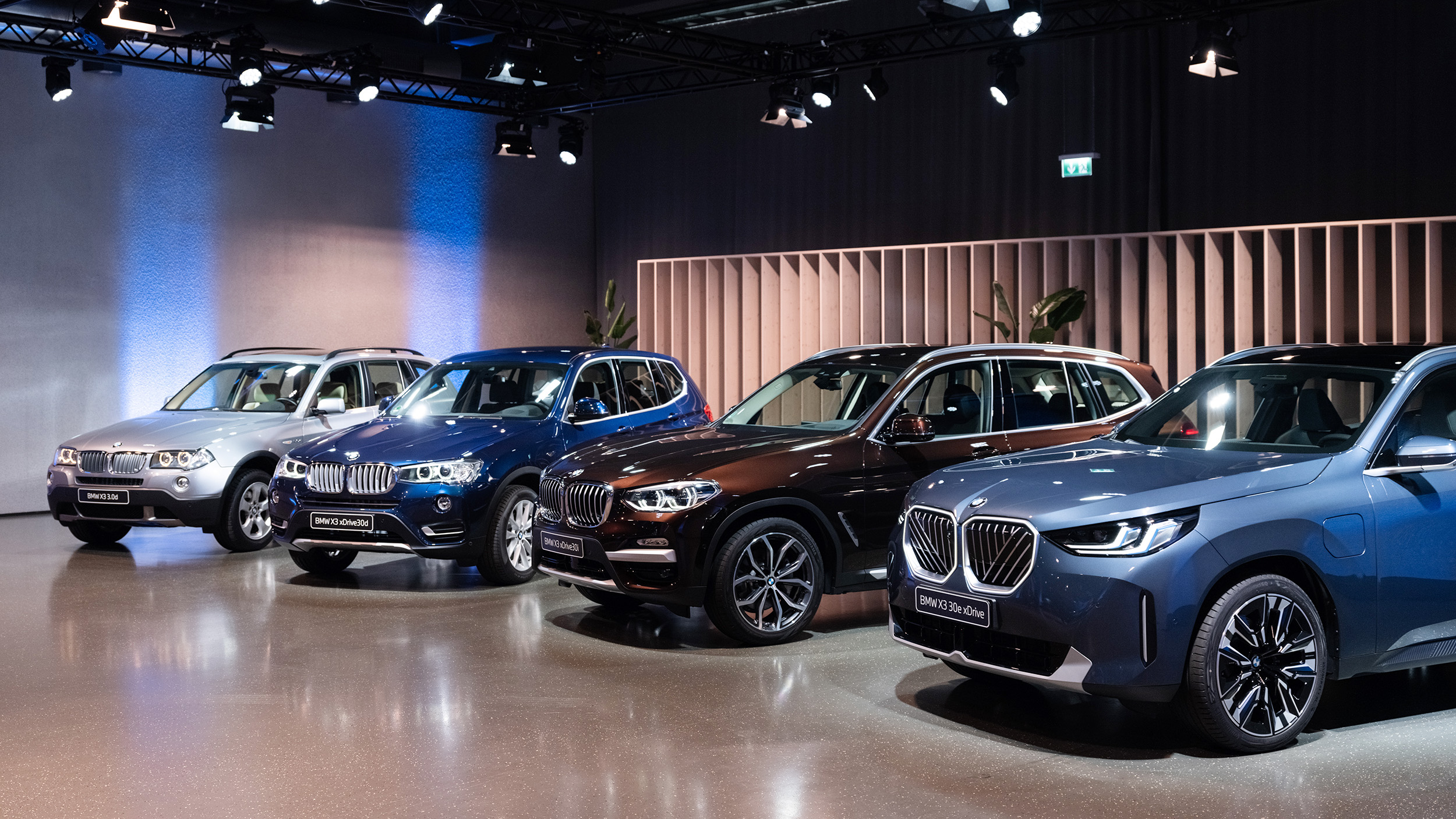Mehrere Generationen eines SUV stehen nach dem Alter aufgereiht in einer Halle, sie werden von Scheinwerfern beleuchtet.