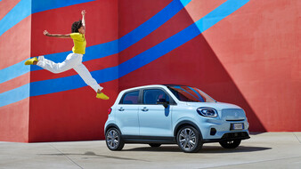 Hellblauer Kleinwagen steht vor einer bunten Wand, eine Frau springt daneben in die Luft