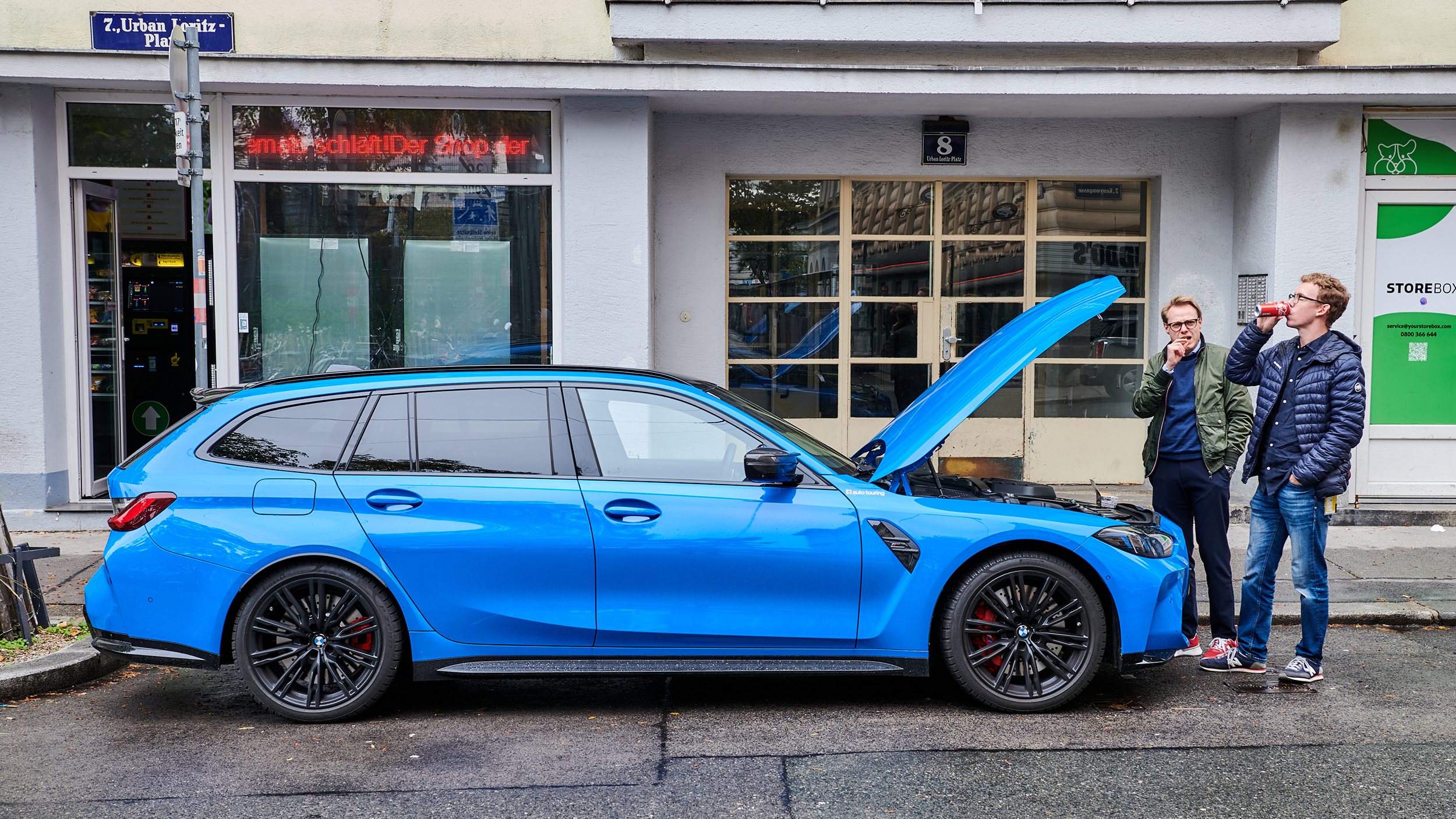 Ein blauer BMW M3 steht mit geöffneter Motorhaube auf einem Parkplatz, zwei Personen stehen neben dem Auto.