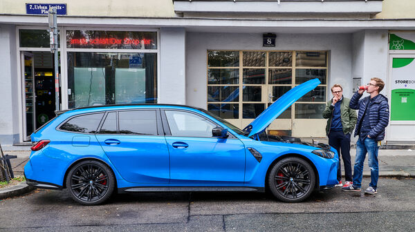 Ein blauer BMW M3 steht mit geöffneter Motorhaube auf einem Parkplatz, zwei Personen stehen neben dem Auto.