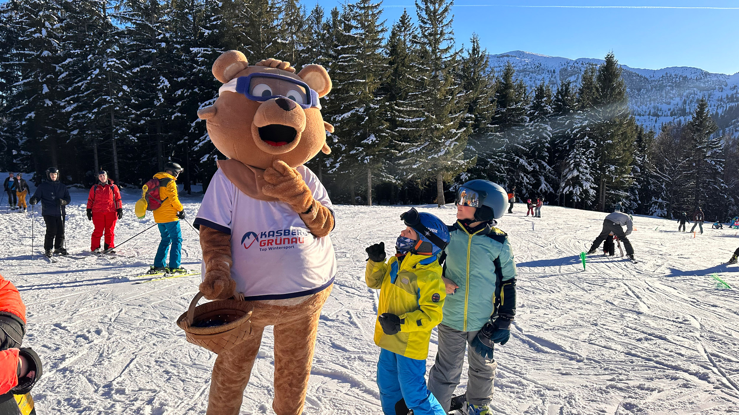 Maskottchen Kasbär Bruno im Bärenkostüm auf der Skipiste am Kasberg. Zwei Kinder stehen gebannt vor ihm. 
