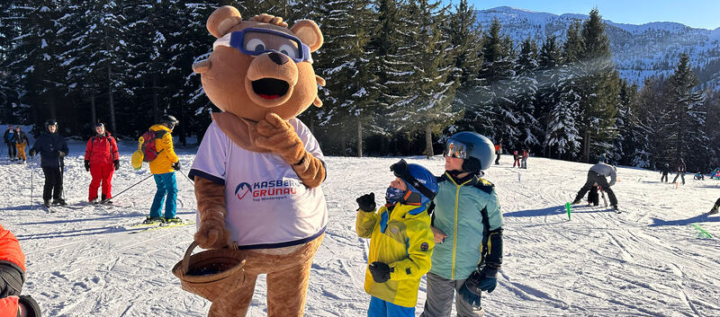 Maskottchen Kasbär Bruno im Bärenkostüm auf der Skipiste am Kasberg. Zwei Kinder stehen gebannt vor ihm.
