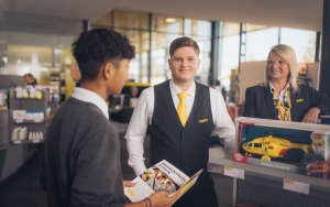 Zwei männliche und eine weibliche Mitarbeiterinnen befinden sich im Schalterbereich eines ÖAMTC Stützpunktes. Alle drei tragen Dienstkleidung (graue Weste, Blazer, Pullover, gelbe Krawatte/Halstuch). 