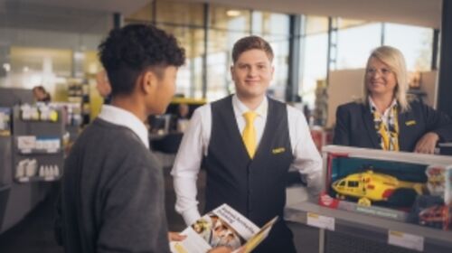 Zwei männliche und eine weibliche Mitarbeiterinnen befinden sich im Schalterbereich eines ÖAMTC Stützpunktes. Alle drei tragen Dienstkleidung (graue Weste, Blazer, Pullover, gelbe Krawatte/Halstuch).