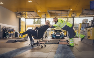 Eine ÖAMTC Kfz-Werkstatt Halle. Im Hintergrund Werkbänke und ein blaues Auto. im Vordergrund schiebt eine Frau in Schalterdienst-Kleidung (dunkelgrauer Blazer und Hose, weiße Bluse) einen Kfz-Technik Kollegen (gelb-graue Jacke und Hose) auf einem Bürostuhl durchs Bild. Beide lachen herzlich.