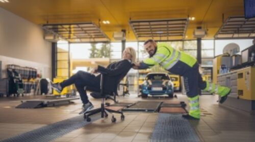 Eine ÖAMTC Kfz-Werkstatt Halle. Im Hintergrund Werkbänke und ein blaues Auto. im Vordergrund schiebt eine Frau in Schalterdienst-Kleidung (dunkelgrauer Blazer und Hose, weiße Bluse) einen Kfz-Technik Kollegen (gelb-graue Jacke und Hose) auf einem Bürostuhl durchs Bild. Beide lachen herzlich.