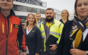 Sechs Personen in unterschiedlicher Dienstkleidung (Flugrettungspilot in roter Jacke, Kfz-Technik Kollege in gelb-grauer Jacke, Kollegin aus dem Reisebüro mit grauer Weste und weißem T-Shirt, Kollegin aus der Fahrtechnik mit schwarzem Polo, Kollege aus dem Büro in Privatkleidung) stehen vor dem Mobilitätszentrum.