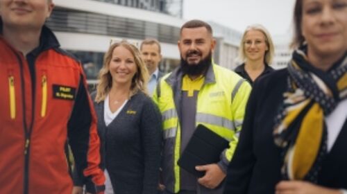Sechs Personen in unterschiedlicher Dienstkleidung (Flugrettungspilot in roter Jacke, Kfz-Technik Kollege in gelb-grauer Jacke, Kollegin aus dem Reisebüro mit grauer Weste und weißem T-Shirt, Kollegin aus der Fahrtechnik mit schwarzem Polo, Kollege aus dem Büro in Privatkleidung) stehen vor dem Mobilitätszentrum.