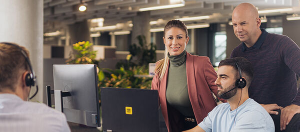 In der ÖAMTC Einsatzzentrale: Drei männliche und zwei weibliche Personen. Drei Personen sitzen mit Headsets am Kopf und haben Bildschirm oder Laptop vor sich. Zwei Personen unterhalten sich stehend.