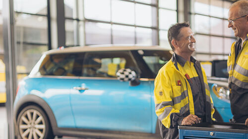 Zwei männliche und eine weibliche Kfz-Technikerin stehen an einer Werkbank in der Kfz-Technik Werkstatt. Im Hintergrund ein blaues Auto auf der Hebebühne. Ein weiterer Kollege kommt im Hintergrund auf die drei Personen zu.
