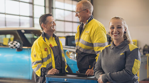 Zwei männliche und eine weibliche Kfz-Technikerin stehen an einer Werkbank in der Kfz-Technik Werkstatt. Im Hintergrund ein blaues Auto auf der Hebebühne. Ein weiterer Kollege kommt im Hintergrund auf die drei Personen zu.