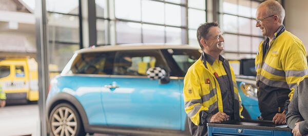 Zwei männliche und eine weibliche Kfz-Technikerin stehen an einer Werkbank in der Kfz-Technik Werkstatt. Im Hintergrund ein blaues Auto auf der Hebebühne. Ein weiterer Kollege kommt im Hintergrund auf die drei Personen zu.