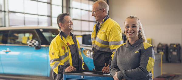 Zwei männliche und eine weibliche Kfz-Technikerin stehen an einer Werkbank in der Kfz-Technik Werkstatt. Im Hintergrund ein blaues Auto auf der Hebebühne. Ein weiterer Kollege kommt im Hintergrund auf die drei Personen zu.