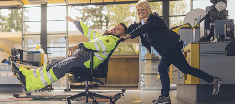 Eine ÖAMTC Kfz-Werkstatt Halle. Im Hintergrund Werkbänke und ein blaues Auto. im Vordergrund schiebt eine Frau in Schalterdienst-Kleidung (dunkelgrauer Blazer und Hose, weiße Bluse) einen Kfz-Technik Kollegen (gelb-graue Jacke und Hose) auf einem Bürostuhl durchs Bild. Beide lachen herzlich.