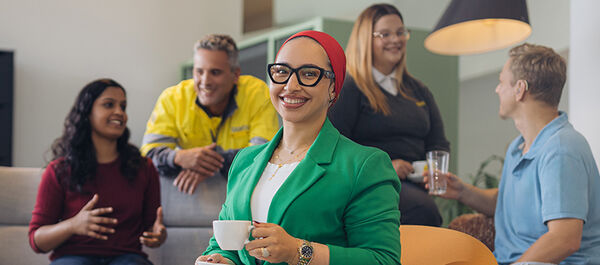 Sieben Personen befinden sich in einem Raum mit Sofa, Stühlen, Lampen, Pflanzen und einem Schrank. Die Einrichtung ist modern, der Raum ist hell und freundlich. Die Frau im Vordergrund lächelt in die Kamera. Sie hat eine Kaffeetasse in der Hand und trägt einen grünen Blazer. Die anderen Personen (vier Männer und zwei Frauen) stehen und sitzen im Raum und unterhalten sich lachend.