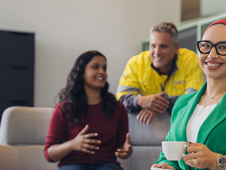 Sieben Personen befinden sich in einem Raum mit Sofa, Stühlen, Lampen, Pflanzen und einem Schrank. Die Einrichtung ist modern, der Raum ist hell und freundlich. Die Frau im Vordergrund lächelt in die Kamera. Sie hat eine Kaffeetasse in der Hand und trägt einen grünen Blazer. Die anderen Personen (vier Männer und zwei Frauen) stehen und sitzen im Raum und unterhalten sich lachend.