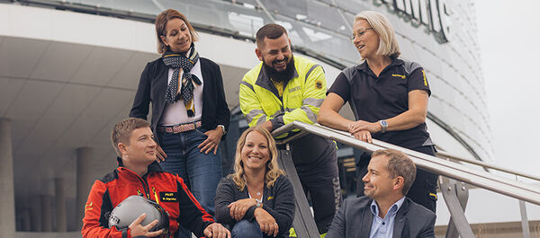 Sechs Personen befinden sich am Treppenaufgang vor dem Mobilitätszentrum im Freien. Drei Personen stehen, drei sitzen. Sie unterhalten sich miteinander und lachen.