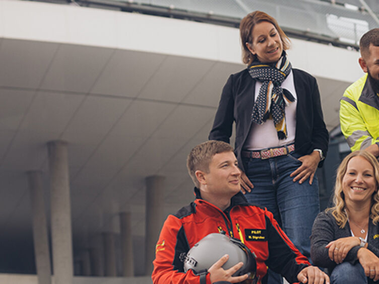 Sechs Personen befinden sich am Treppenaufgang vor dem Mobilitätszentrum im Freien. Drei Personen stehen, drei sitzen. Sie unterhalten sich miteinander und lachen.