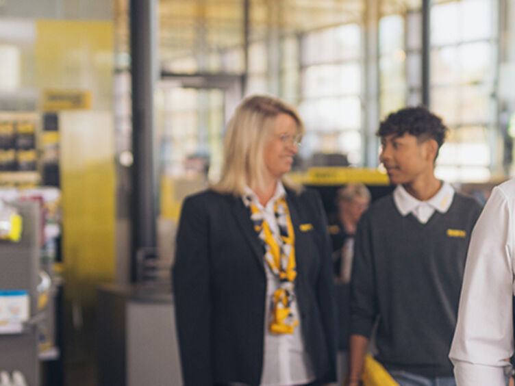 Zwei männliche und eine weibliche Mitarbeiterinnen befinden sich im Schalterbereich eines ÖAMTC Stützpunktes. Alle drei tragen Dienstkleidung (graue Weste, Blazer, Pullover, gelbe Krawatte/Halstuch). Ein Kollege steht im Vordergrund und lehnt sich lächelnd an ein Regal. Die anderen beiden unterhalten sich im Hintergrund.