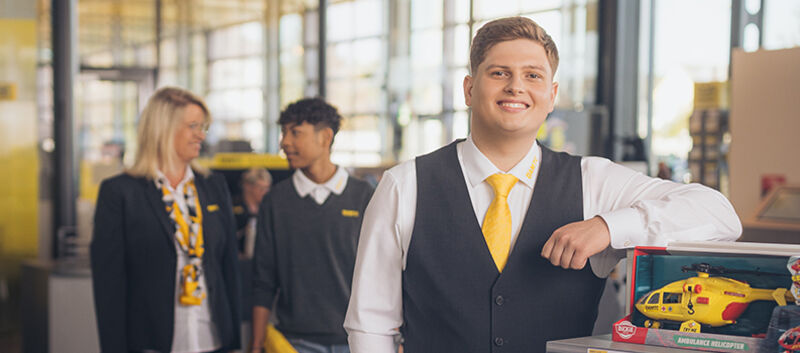 Zwei männliche und eine weibliche Mitarbeiterinnen befinden sich im Schalterbereich eines ÖAMTC Stützpunktes. Alle drei tragen Dienstkleidung (graue Weste, Blazer, Pullover, gelbe Krawatte/Halstuch). Ein Kollege steht im Vordergrund und lehnt sich lächelnd an ein Regal. Die anderen beiden unterhalten sich im Hintergrund.