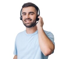 Junger Mann mit kurzem schwarzen Haar und Bart lächelt in die Kamera. Er trägt ein Headset und greift mit seiner linken Hand an sein linkes Ohr. Er hat ein hellblaues T-Shirt an.