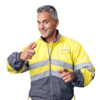 Mann mit grauen Haaren in Kfz-Techniker Dienstkleidung (grau-gelbe Jacke, graue Hose) zeigt lächelnd in die Kamera.